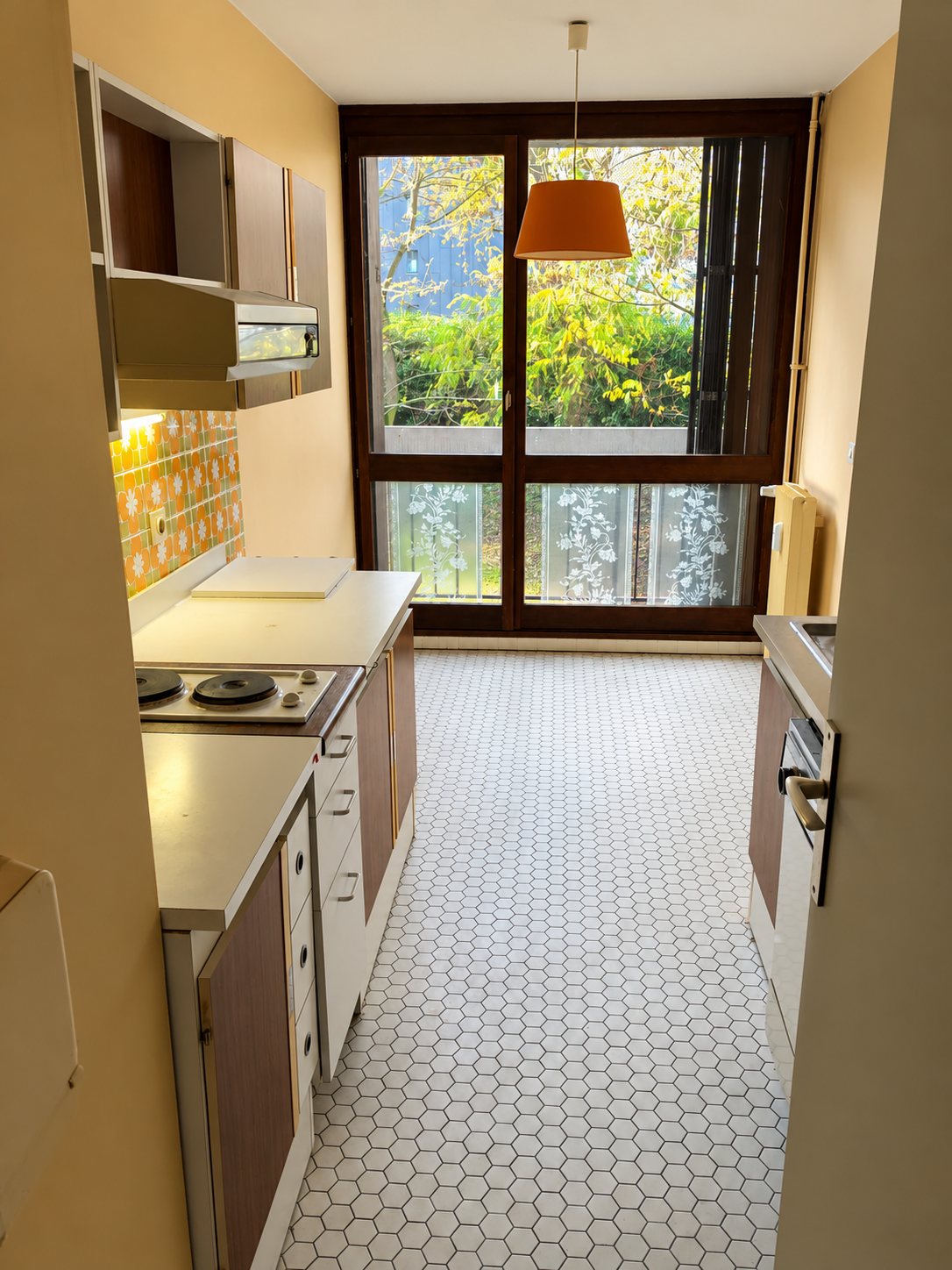 Cuisine étroite et datée avec plaques électriques, meubles beiges, carrelage hexagonal au sol et grande fenêtre donnant sur des arbres.
