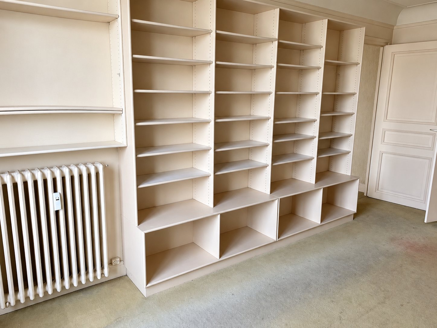 Une grande bibliothèque en bois clair à étagères réglables occupe le mur d'une pièce vide, à côté d'un radiateur blanc et d'une porte moulurée.