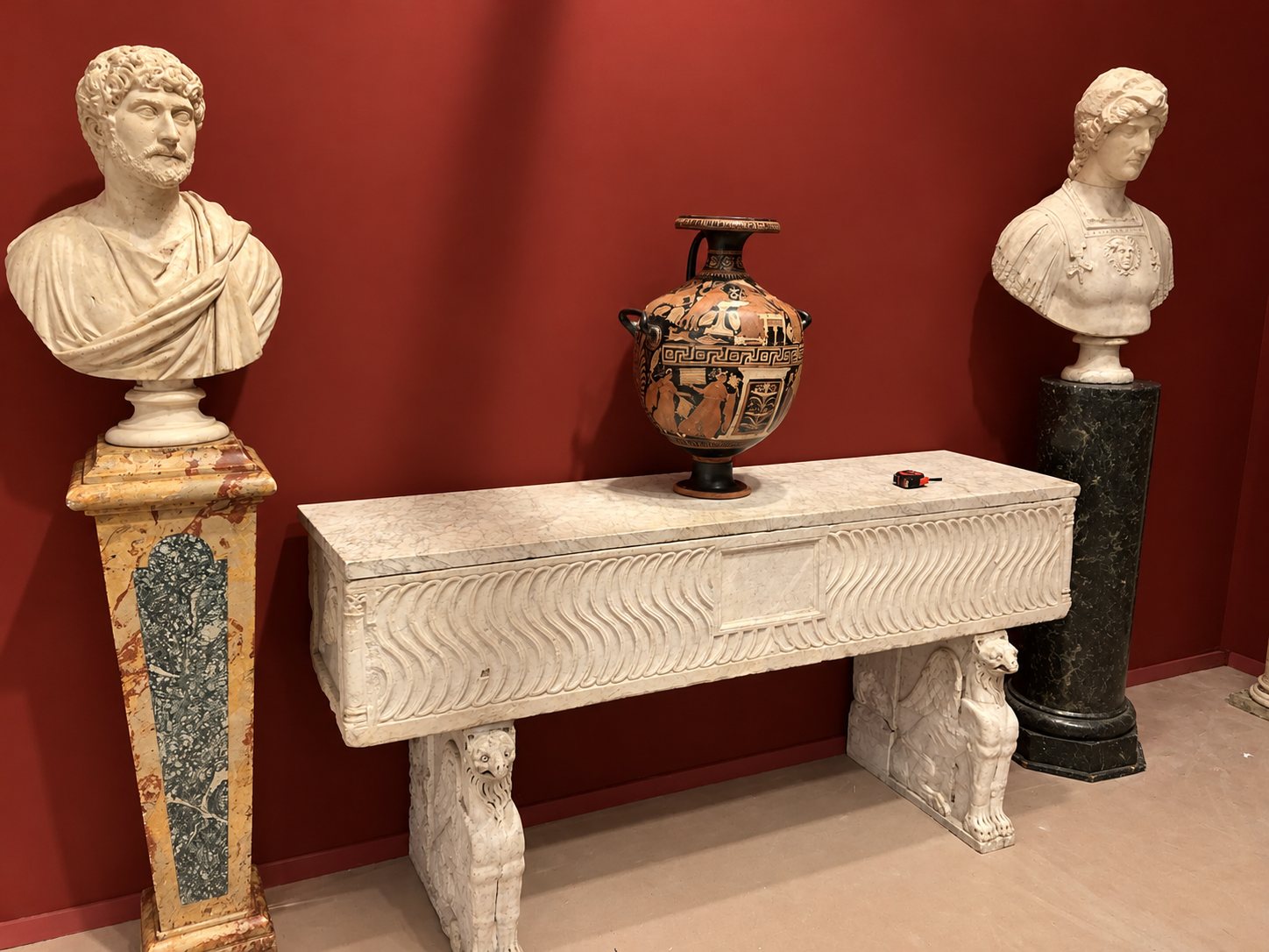 Deux bustes en marbre sur des piédestaux encadrent une console en marbre sculptée aux pieds de griffons, surmontée d'un vase grec, devant un mur rouge.