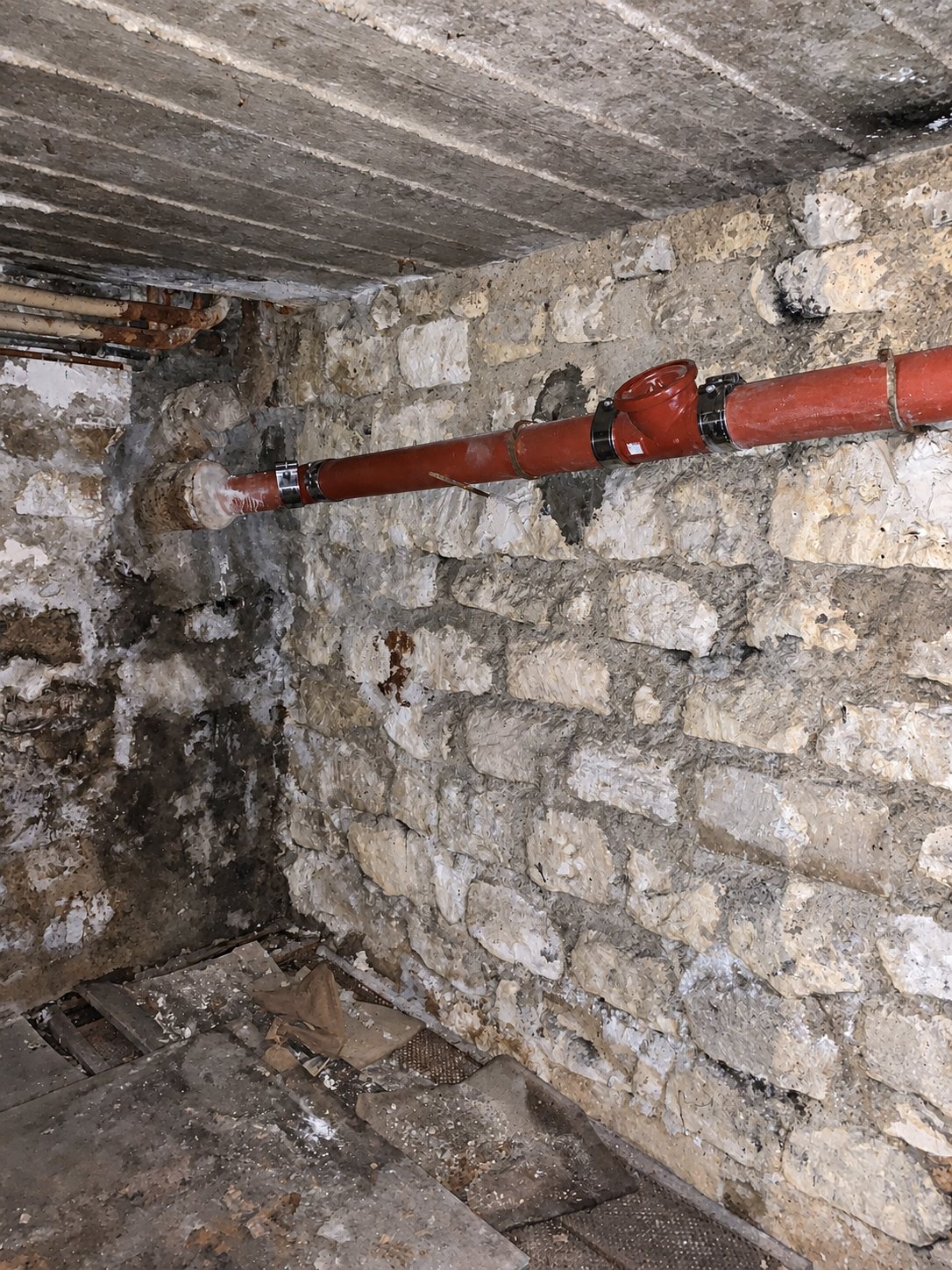 Une canalisation d'évacuation rouge est fixée à un mur de pierres apparentes dans une cave sombre au plafond de planches.