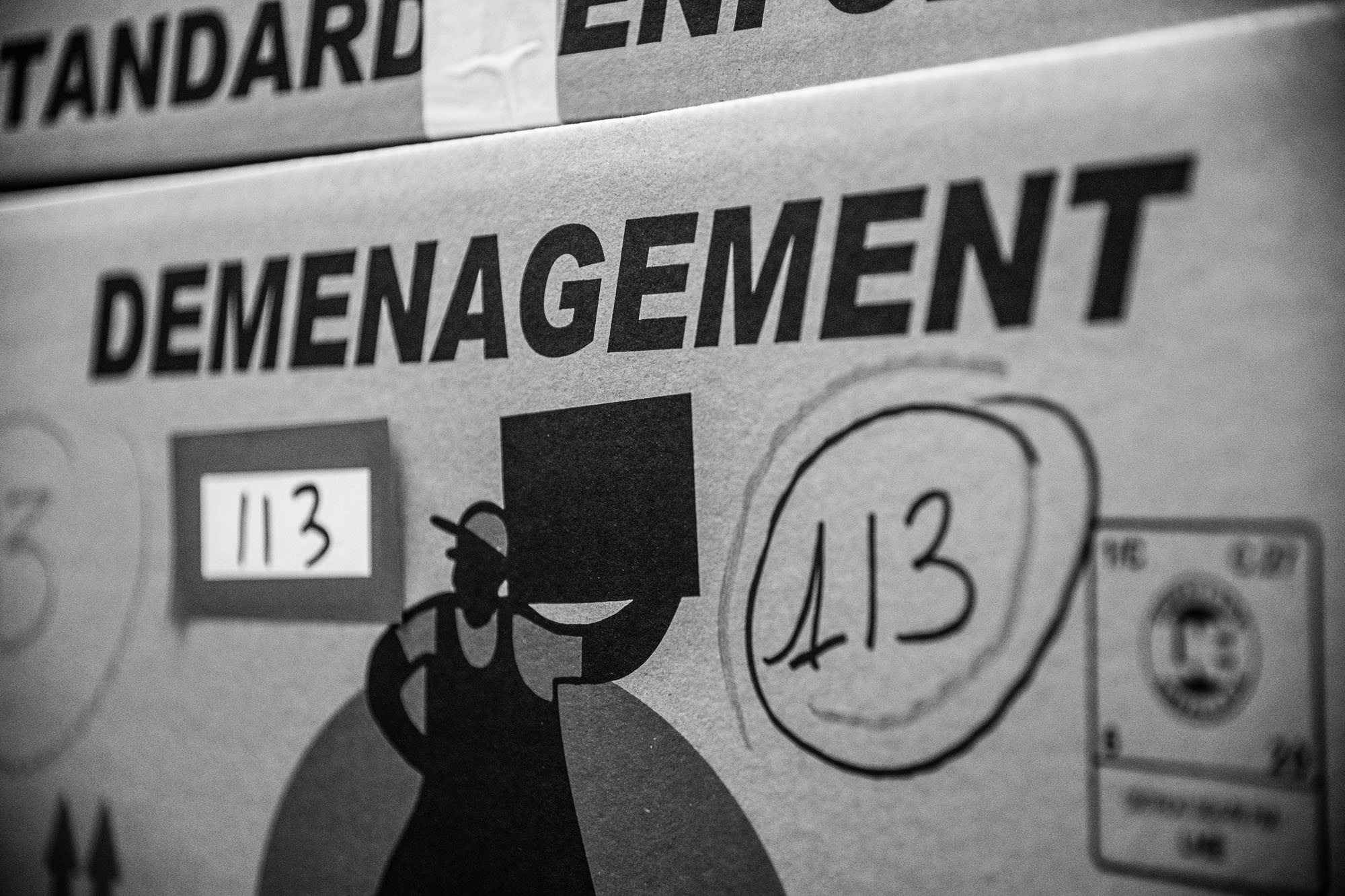 Gros plan en noir et blanc sur un carton de déménagement portant le numéro 113, écrit à la main et sur une étiquette, avec le dessin d'un déménageur portant une caisse.