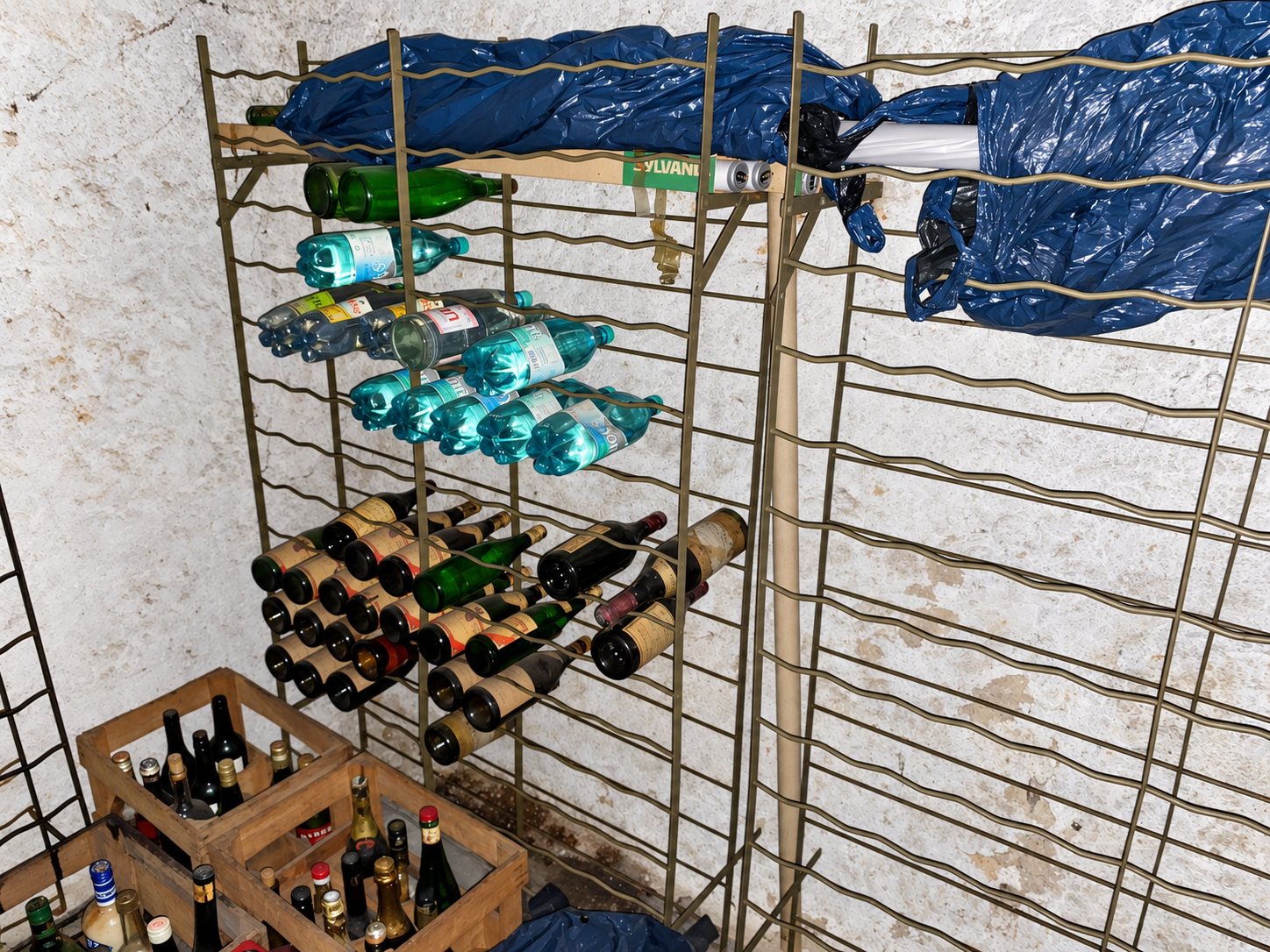 Dans une cave aux murs blanchis, des casiers métalliques contiennent des bouteilles de vin et d'eau, avec des caisses en bois remplies de bouteilles au sol.