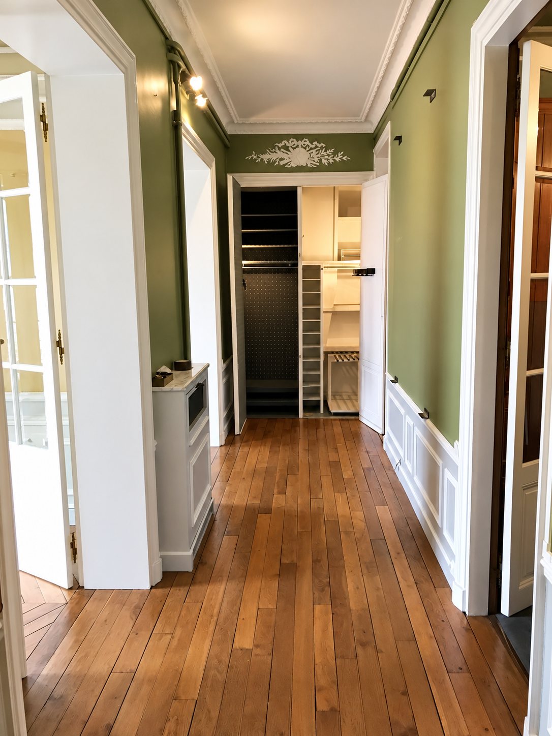 Un couloir d'appartement ancien aux murs vert clair, avec un parquet en bois, des boiseries blanches et un grand placard ouvert au fond.