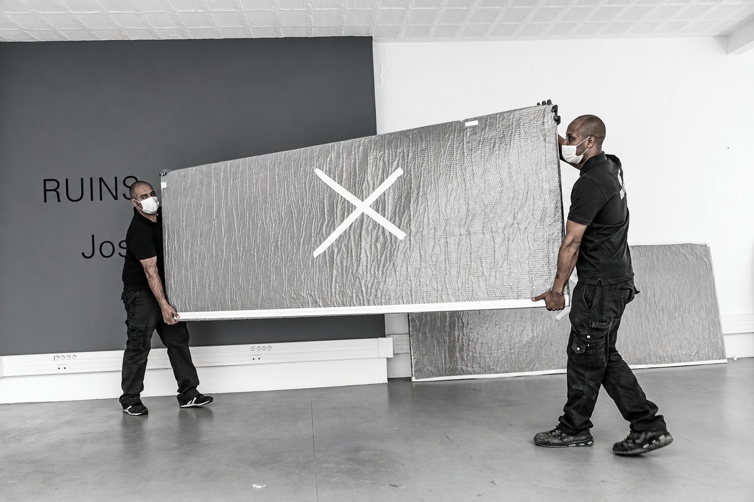 Deux hommes masqués en vêtements noirs transportent un grand panneau emballé marqué d'une croix devant un mur portant le mot RUINS, dans une salle d'exposition.
