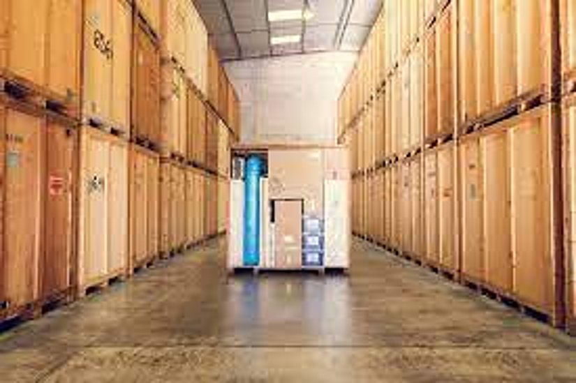 Une allée d'entrepôt éclairée bordée de hautes étagères remplies de grandes caisses en bois, avec une palette chargée au fond.