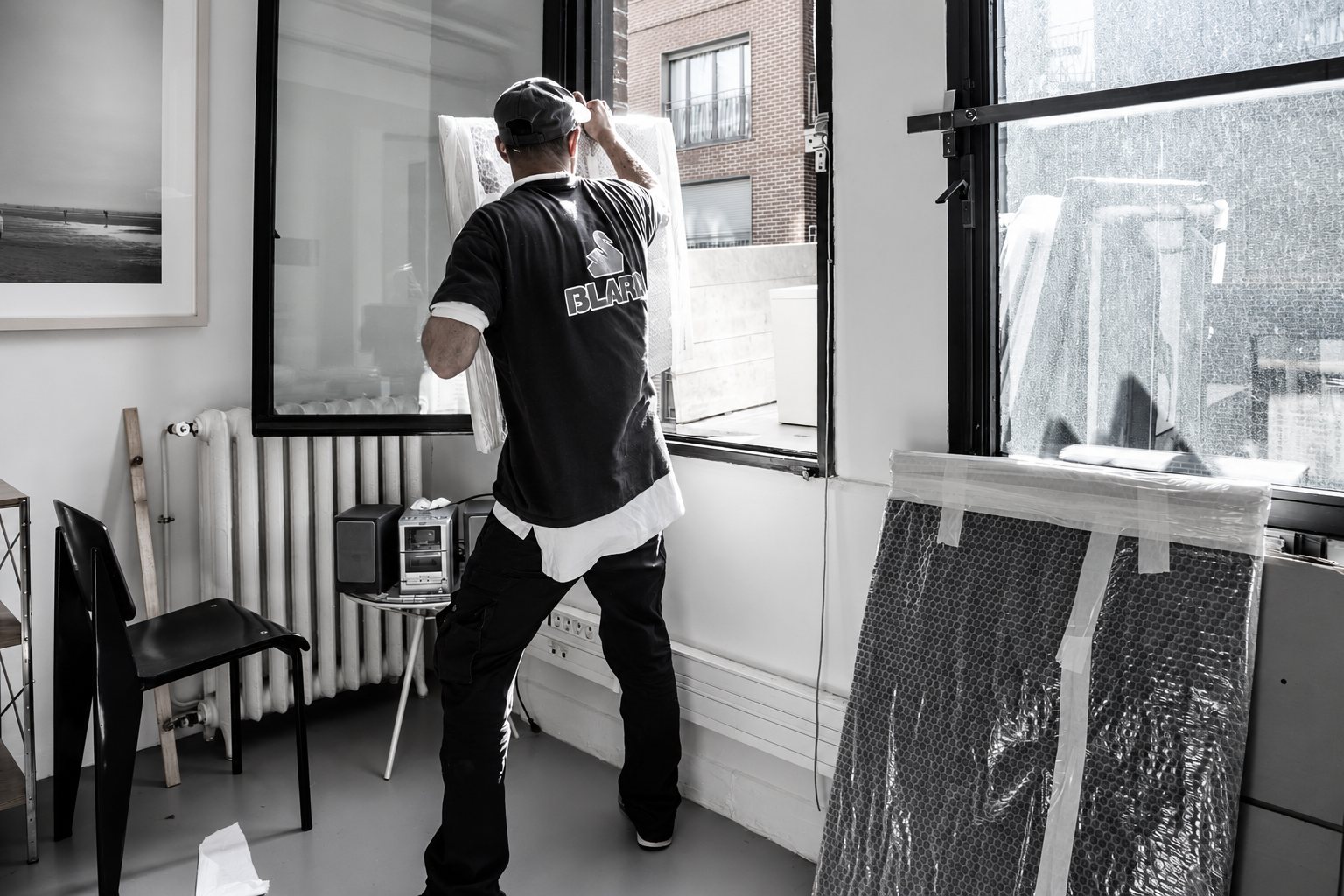 Un homme de dos, en t-shirt sombre, manipule une grande œuvre protégée par du plastique dans une pièce lumineuse, à côté d'un radiateur et d'une chaise noire.