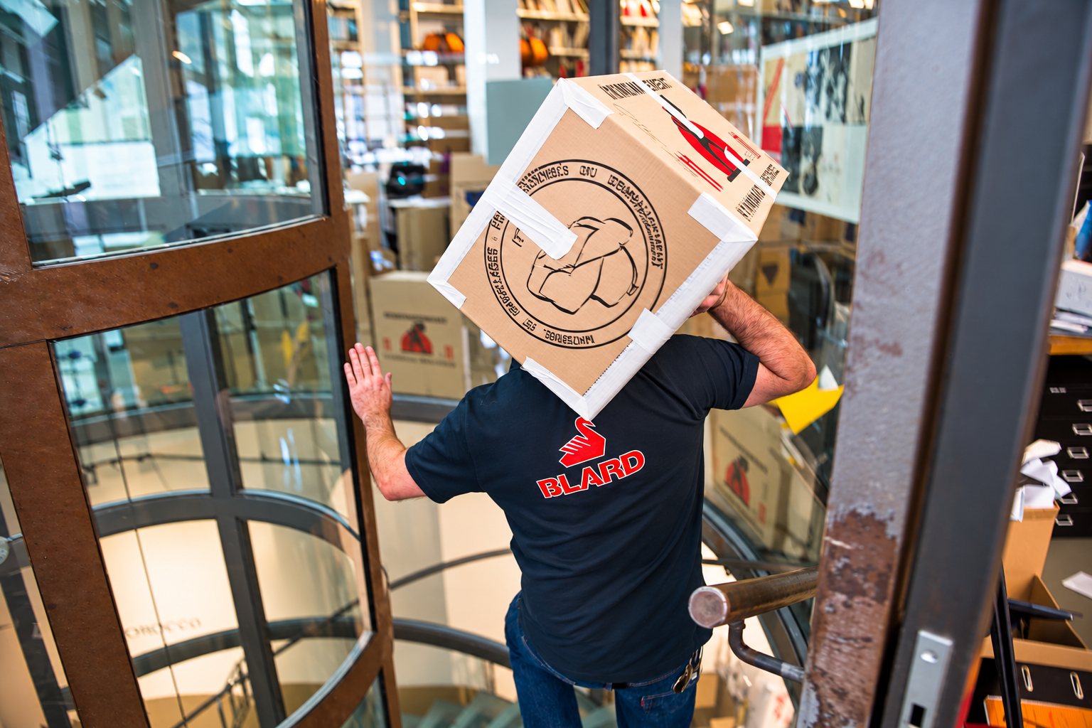 Un homme en t-shirt bleu marine floqué Blard porte un grand carton sur l'épaule en passant une porte tournante vitrée.