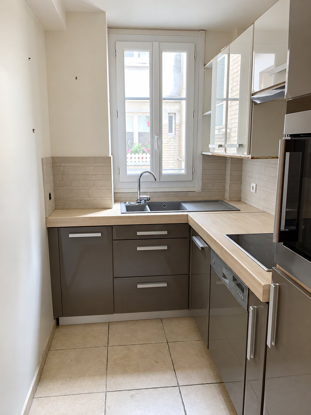 Petite cuisine en L aux meubles gris brillant, plan de travail en bois clair, évier encastré sous une fenêtre et carrelage beige au sol.