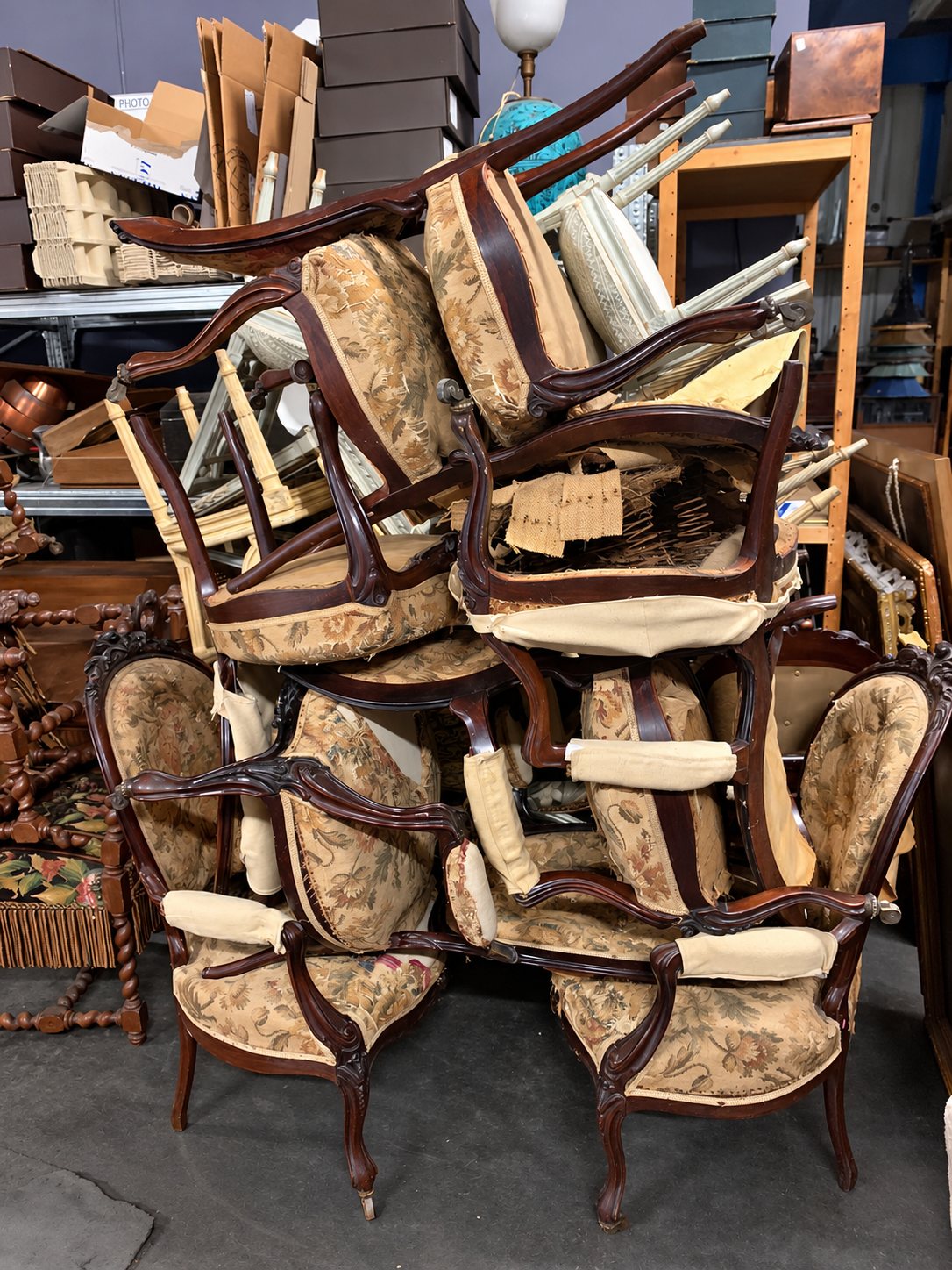 Un empilement de fauteuils anciens en bois sombre, tapissés de tissu à motifs floraux et en cours de restauration, dans un atelier encombré.