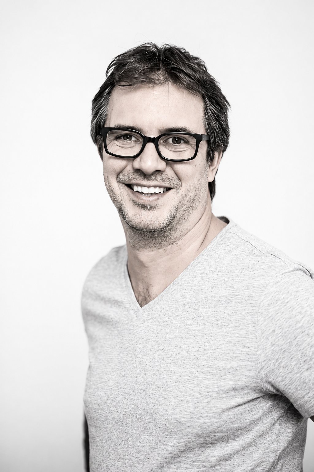 Portrait en noir et blanc d'un homme souriant, portant des lunettes à monture épaisse et un t-shirt à col en V, devant un fond uni clair.