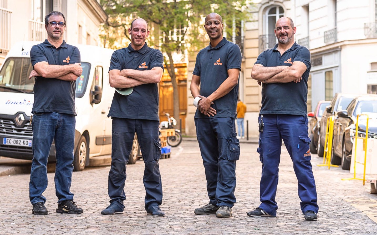 Quatre hommes en polo et pantalon de travail posent les bras croisés dans une rue pavée, devant une camionnette blanche garée.