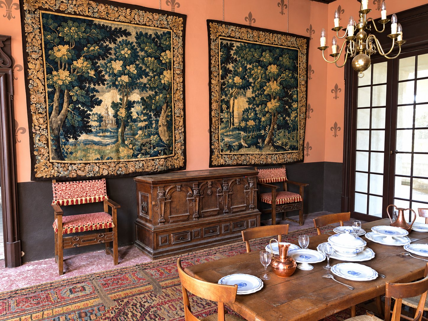 Salle à manger ancienne aux murs ornés de fleurs de lys et de deux grandes tapisseries, avec un coffre en bois sculpté, des fauteuils et une table dressée de vaisselle bleue et blanche.