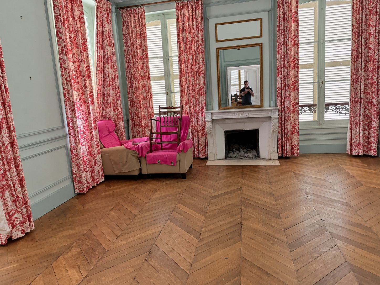 Un grand salon de style ancien avec parquet en chevrons, cheminée en marbre blanc, miroir, rideaux à motifs rouges et fauteuils recouverts de tissu rose.