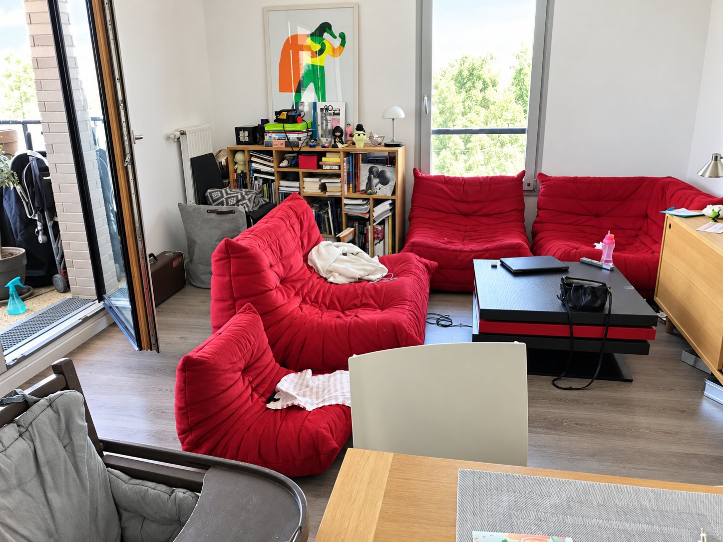 Un salon avec des éléments de canapé rouge en tissu matelassé autour d'une table basse noire, une bibliothèque en bois et une porte-fenêtre ouverte sur un balcon.