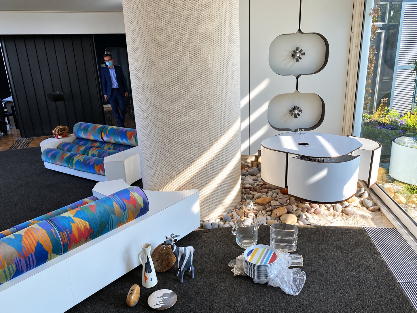 Un salon de style rétro avec des canapés blancs aux coussins multicolores, une table basse ronde, des galets au sol et des objets en céramique posés sur la moquette sombre.
