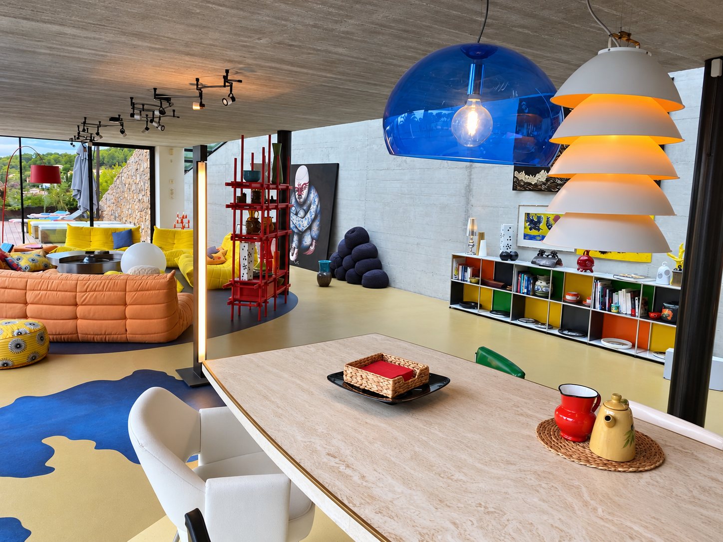 Vaste pièce de vie au plafond de béton avec canapés orange et jaunes, table en travertin au premier plan, suspensions bleue et blanches et bibliothèque colorée.
