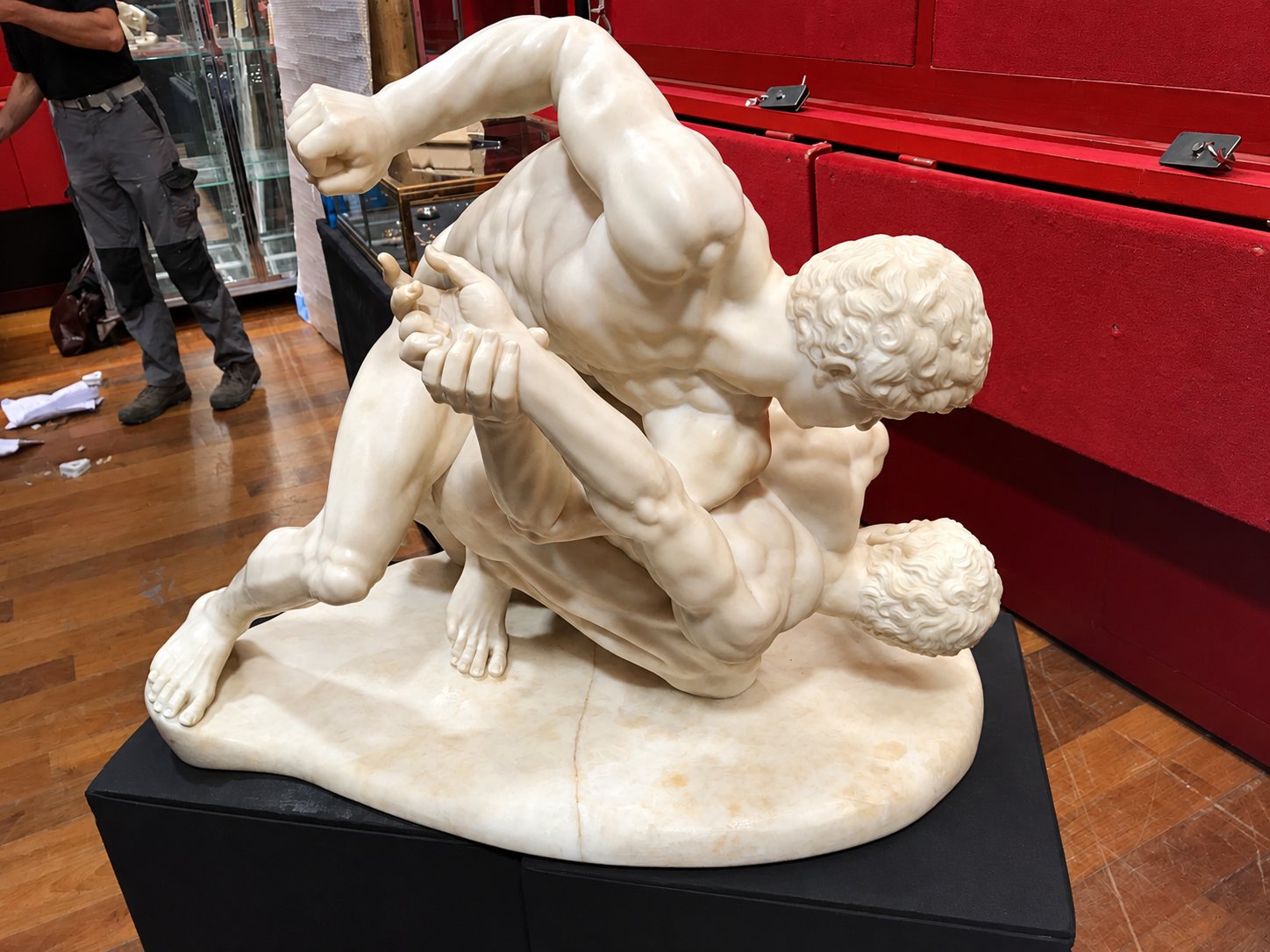 Une sculpture en marbre blanc représentant deux lutteurs musclés enlacés, posée sur un socle noir dans une salle au parquet et aux murs rouges.
