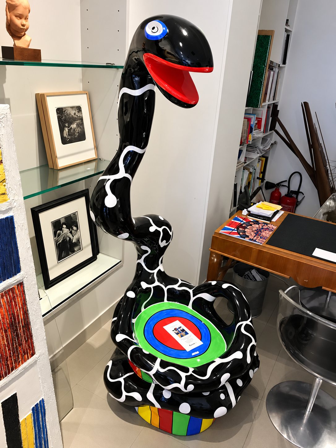 Une grande sculpture de serpent noir à motifs blancs, à la gueule rouge et au socle multicolore, se dresse dans une galerie entre des étagères en verre, des cadres et un bureau en bois.