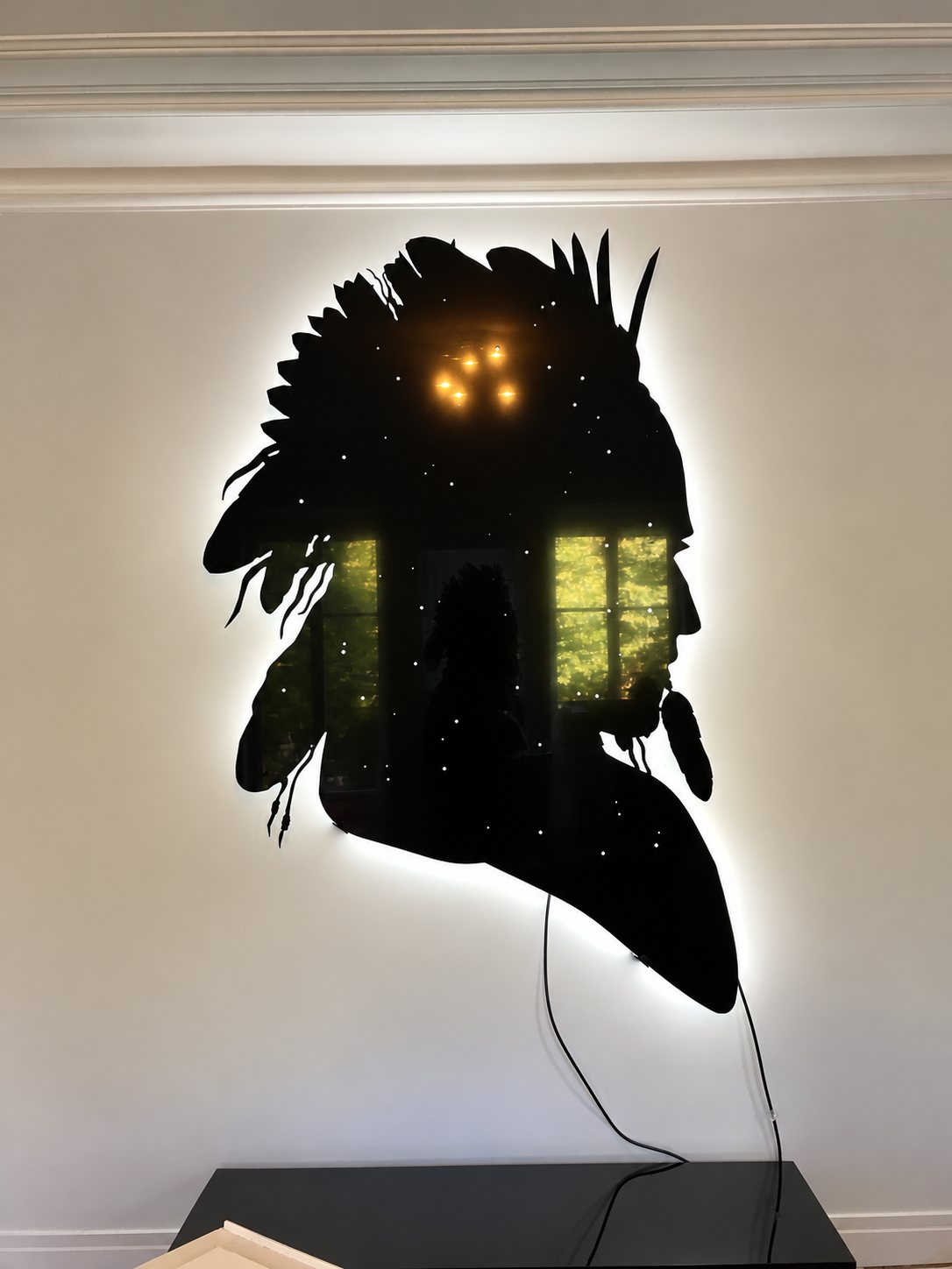 Une grande silhouette noire de profil coiffé de plumes est fixée au mur, rétroéclairée par une lumière blanche et percée de petits points lumineux, avec des câbles qui pendent au-dessus d'une console sombre.
