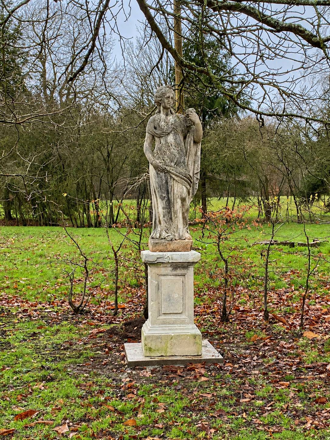 Une statue en pierre patinée représentant une figure drapée se dresse sur un piédestal carré au milieu d'une pelouse parsemée de feuilles mortes, entourée d'arbres nus.