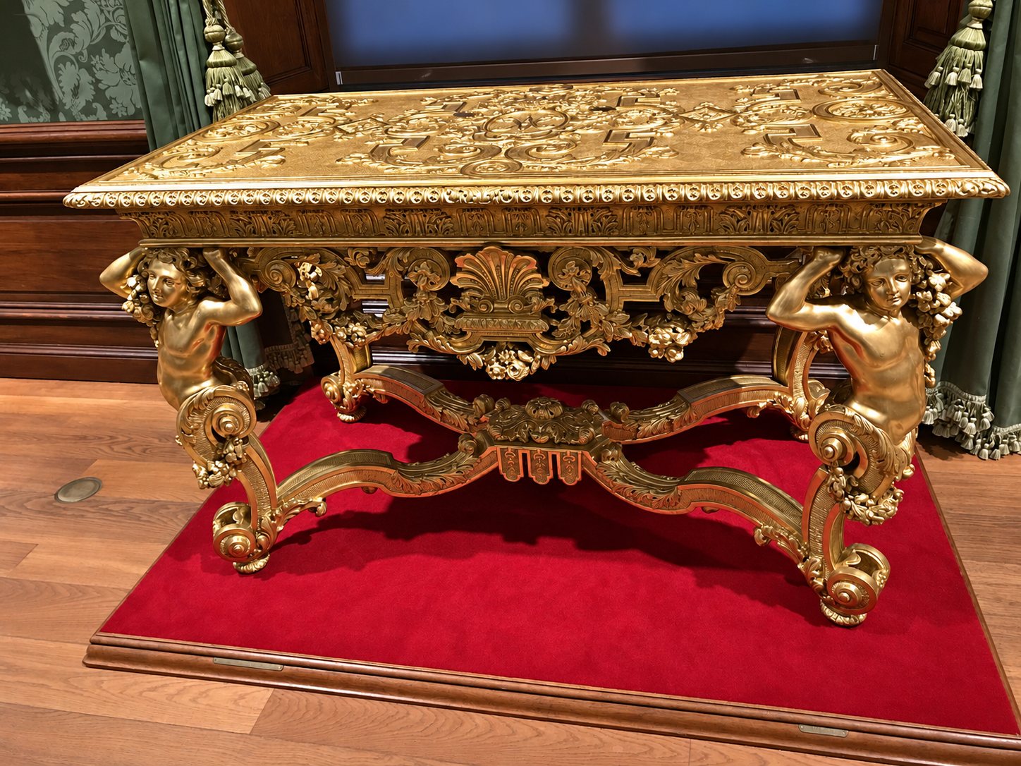 Une table baroque en bois doré richement sculptée, soutenue par deux putti, repose sur une estrade recouverte de velours rouge dans une salle de musée.