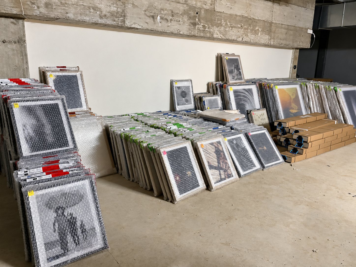 Des dizaines de cadres protégés par du papier bulle sont alignés contre un mur blanc dans une salle de stockage en béton, à côté de piles de cartons.