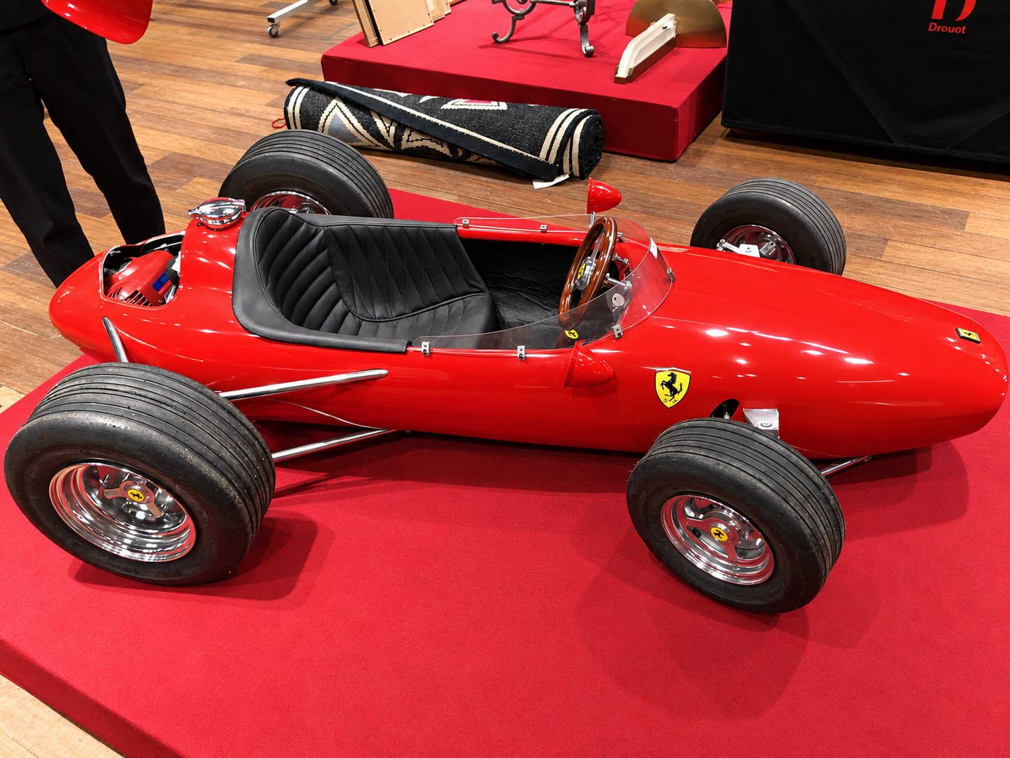 Une petite voiture de course rouge de style Ferrari, avec siège en cuir noir et volant en bois, est présentée sur un podium rouge dans une salle de vente.